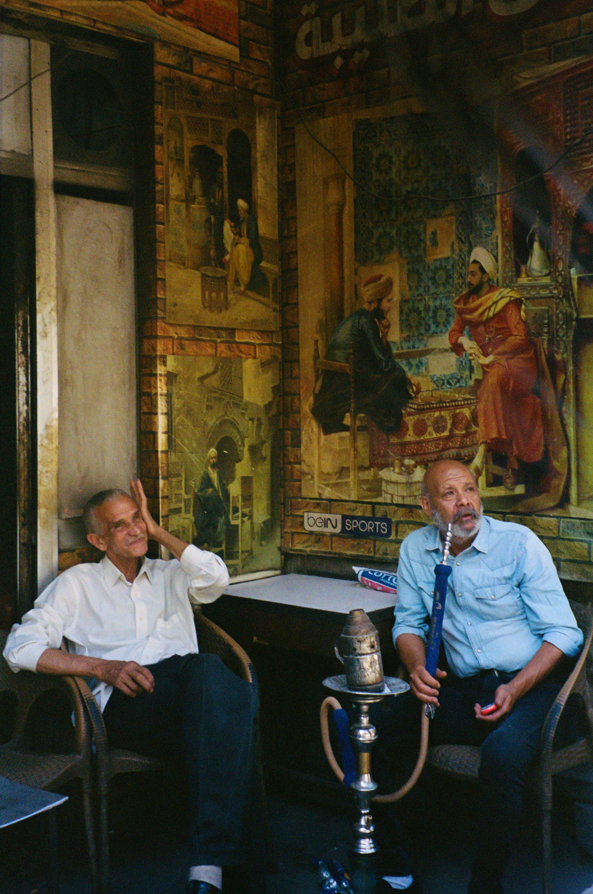 Two men sitting with a shisha pipe beneath a painted mural in a Cairo cafe, Egypt, film photography by Loud Etudes